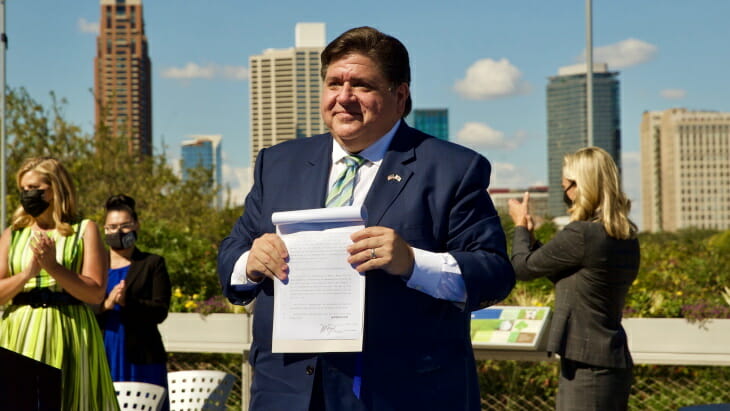 El gobernador Pritzker sostiene una legislación energética recientemente firmada frente a un soleado horizonte de Chicago, con una multitud de espectadores enmascarados a la izquierda de la imagen.